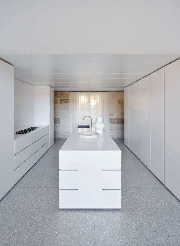 A bright, minimalist galley kitchen viewed straight toward a central island. The kitchen features extensive seamless white cabinetry, integrated drawers, a white island with a dark gooseneck tap and basin, a concealed cooktop on the left, a grey speckled floor, a white panelled ceiling, and a textured natural-stone feature wall behind sheer curtains.
