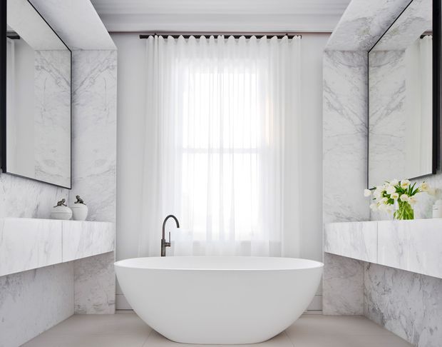 Bright, symmetrical luxury bathroom featuring a freestanding oval bathtub centered beneath a tall sheer-curtained window. Veined white marble-look wall panels and matching floating vanity surfaces flank the bath, with large mirrored cabinets, a dark minimalist faucet, white vessel basins, and understated floral styling. The space has a clean, refined, high-end contemporary appearance.