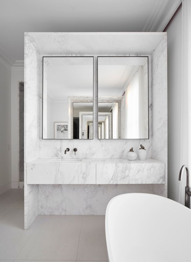 Bright, refined bathroom featuring a freestanding white bathtub, a wall-mounted double vanity, and large rectangular mirrors set within a dramatic white marble surround. The space has pale tiled flooring, white walls with classic moulded detailing, brushed dark-metal tapware, and strong mirror reflections creating depth.