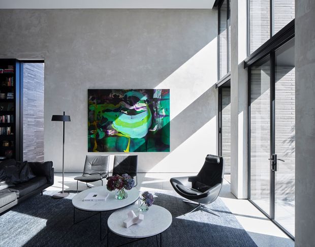 A spacious contemporary living room with a double-height feel, polished concrete-style walls, expansive black-framed floor-to-ceiling glazing, and strong geometric sunlight. The room features a dark sectional sofa, two black leather lounge chairs, round white coffee tables, a black floor lamp, a large abstract artwork, and a dark area rug. An exterior brick wall is visible through the glazing.