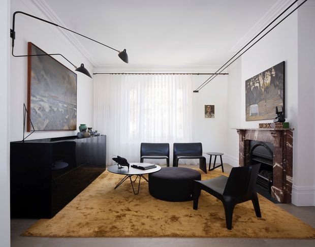 A refined living room with a symmetrical, light-filled layout. The room features white walls and ornate ceiling cornices, a dark marble fireplace surround, a large black built-in sideboard, mid-century-style black leather lounge chairs, a low round coffee table, a broad tan rug, and sculptural black linear pendant lighting. A large artwork and sheer curtains add softness while the fireplace provides a traditional architectural focal point.