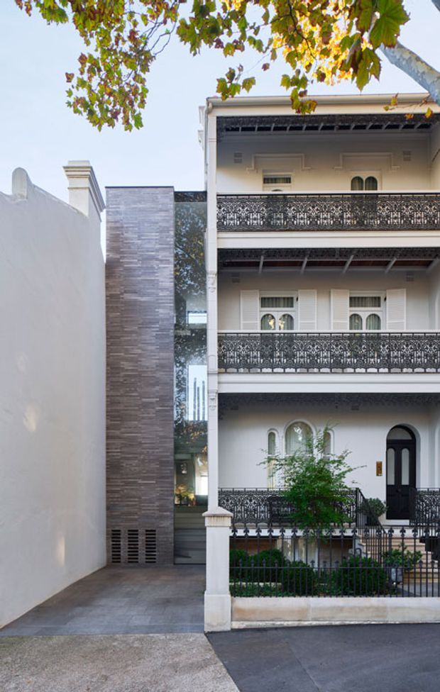 Street-facing view of a narrow, multi-storey heritage-style residence with a white rendered facade, ornate dark metal balcony balustrades, decorative mouldings, shuttered windows, arched openings and a black arched front door. A tall contemporary stone-clad wall and glazed vertical section form an adjoining entry or side element. The property has a small planted front garden behind metal fencing and a paved driveway or forecourt. Tree branches frame the upper edge of the image.