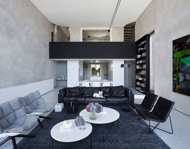 A sophisticated double-height open-plan living room with exposed textured grey walls, a dark suspended mezzanine or volume overhead, and a large black built-in shelving unit. The room contains a black leather sofa, pale blue lounge chairs, black leather armchairs, round white coffee tables, and a dark rug. A dining area and compact kitchen are visible beyond, with contemporary lighting, artwork, and streamlined built-in cabinetry.