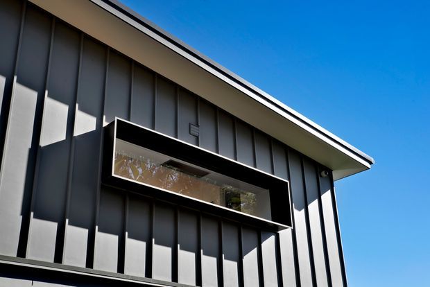 Upward-looking exterior view of a modern building facade with dark vertical panel cladding, a sharply sloping roof edge, and a projecting rectangular balcony or window box with dark framing and glazing. Bright blue sky is visible around the building.