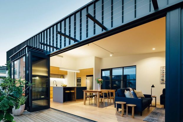 A contemporary open-plan apartment interior opening onto a timber deck. The space combines a compact kitchen with a dark island and timber cabinetry, a dining table, and a living area with a deep blue sofa. Full-height black-framed glazing and a projecting upper-level structure create a strong indoor-outdoor connection, while plants and warm timber finishes soften the modern interior.