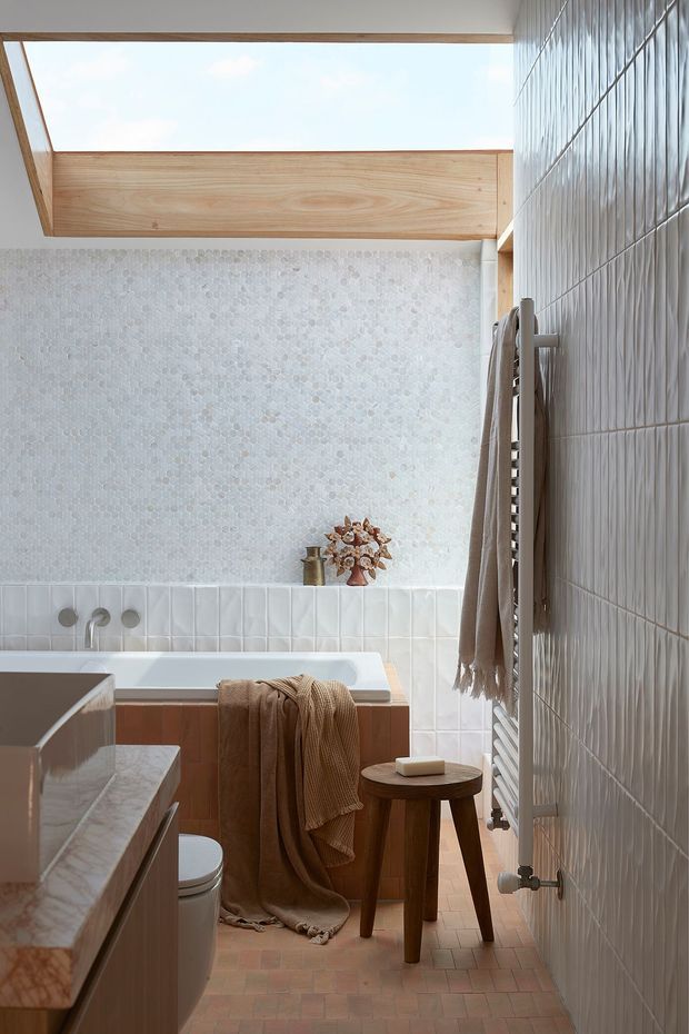 A softly lit bathroom with a freestanding bathtub beneath a large skylight, surrounded by pale mosaic and vertically ridged wall tiles. Warm timber framing, terracotta floor tiles, a timber vanity, brass fixtures, a towel radiator, and a small wooden stool create a warm, refined atmosphere.