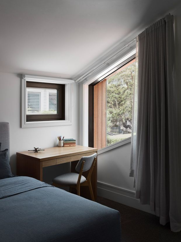 A compact, softly lit bedroom with a neatly made blue-grey bed, a simple timber writing desk, and a mid-century-inspired upholstered chair. White walls and detailed window trims frame a small square window and a larger timber-lined window overlooking greenery. Full-height grey curtains and dark carpet create a calm, muted atmosphere.