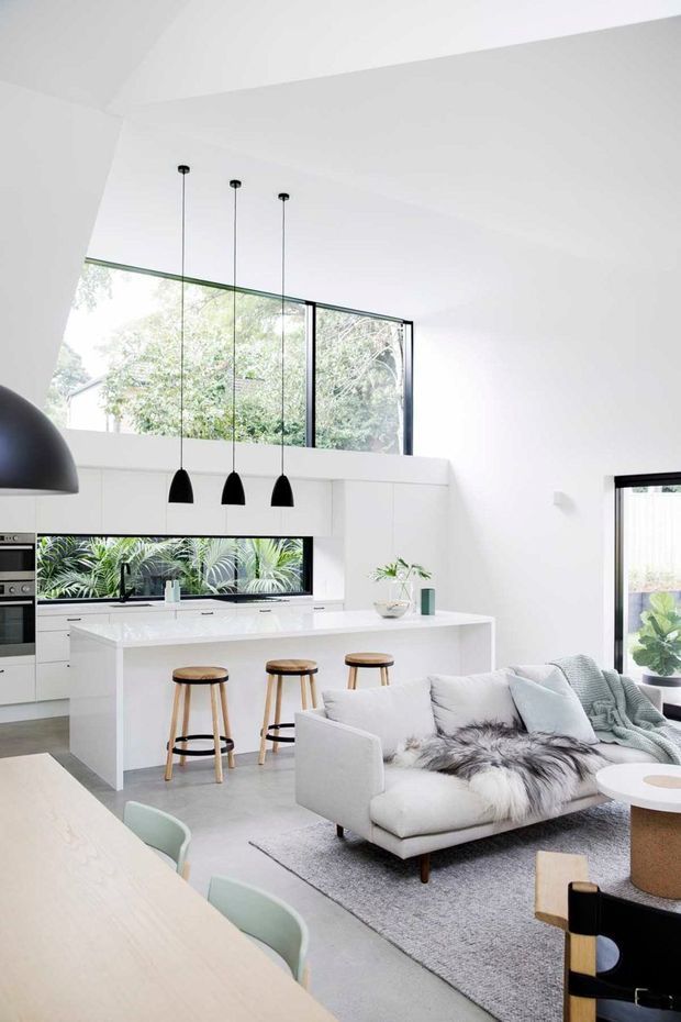 Bright open-plan interior combining a minimalist white kitchen with island seating and a contemporary living area. The room features high ceilings, upper clerestory-style glazing, large black-framed windows and doors, white cabinetry and benchtops, pale flooring, a light sofa, pendant lights, and a dining table in the foreground.