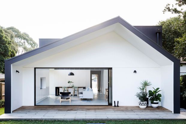 Front exterior view of a contemporary single-storey gable-roof extension or pavilion. The white rendered facade has a sharply pitched gable, dark roof edging and trim, broad black-framed glazing, and a timber deck with potted plants. A bright, minimalist living area and dining space are visible through the glazed opening.