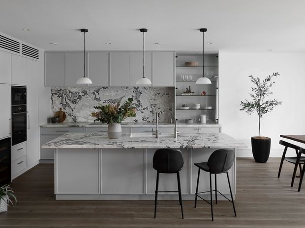 A sophisticated contemporary kitchen with a large marble-patterned island, pale grey flat-panel cabinetry, integrated appliances, open shelving, and three pendant lights. Black bar stools line the island, while a dining table and potted tree occupy the adjacent area. Dark timber flooring and a dramatic veined stone splashback add contrast to the otherwise light, refined interior.