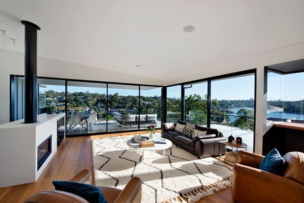 Bright contemporary open-plan living room with extensive black-framed floor-to-ceiling glazing overlooking a leafy waterfront setting. The room features warm timber flooring, a white fireplace surround with a tall cylindrical black flue, a dark leather sofa, leather armchairs, a round coffee table, and a large geometric cream rug. A dining area and kitchen or serving zone are visible beyond the living space.