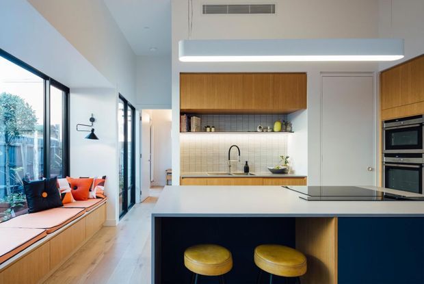 Bright open-plan kitchen and living area with a streamlined island, integrated cooking appliances, warm timber cabinetry, pale tiled splashback, and a built-in window seat beside full-height glazing. Two mustard-yellow stools provide a restrained accent against the dark island panels.