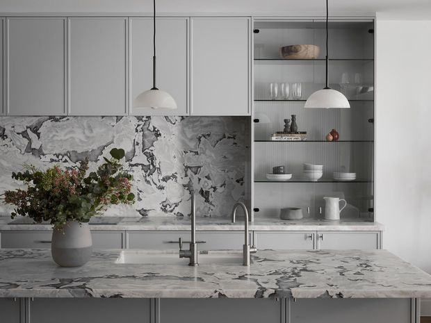 A refined contemporary kitchen viewed straight on, featuring pale grey-blue framed cabinetry, a large island with a dramatic grey and white veined stone surface, matching abstract-veined splashback, open glass shelving with curated tableware, and two simple white pendant lights. A sculptural vase with greenery sits on the island, adding an organic accent to the otherwise cool, monochromatic scheme.
