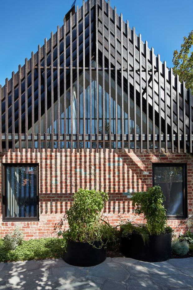 Exterior view of a distinctive gabled building facade. A red brick lower wall with dark-framed windows supports a steep, sharply peaked roof form enclosed by extensive vertical dark metal fins and glazing. The facade creates strong linear shadows, with planted black raised containers and paving in the foreground under clear daylight.