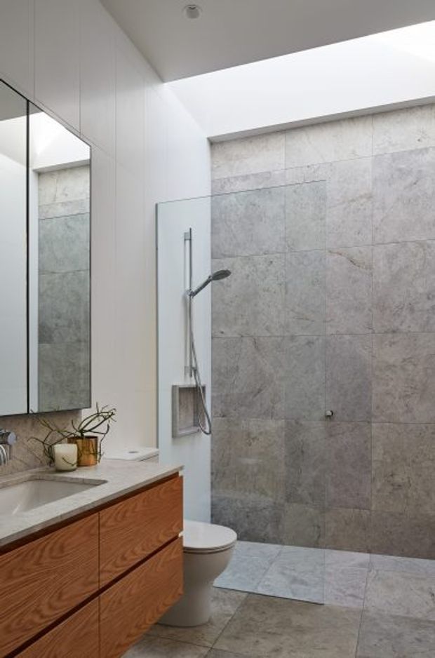 Bright contemporary bathroom with a large walk-in shower, full-height grey stone-look wall tiling, matching floor tiles, floating warm timber vanity with a pale stone benchtop, mirrored cabinetry, wall-mounted shower fixtures, and a skylight or clerestory opening providing natural light.