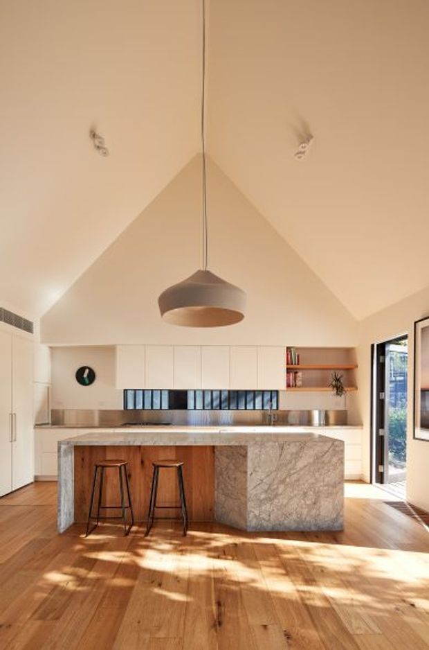 A bright contemporary kitchen viewed from the living area, featuring a large marble-clad island with timber cabinetry and two bar stools, white full-height cabinetry, a stainless-steel worktop and splashback, open timber shelving, and warm timber flooring. A vaulted ceiling with a large sculptural pendant adds vertical emphasis, while sunlight enters through the open glazed door.