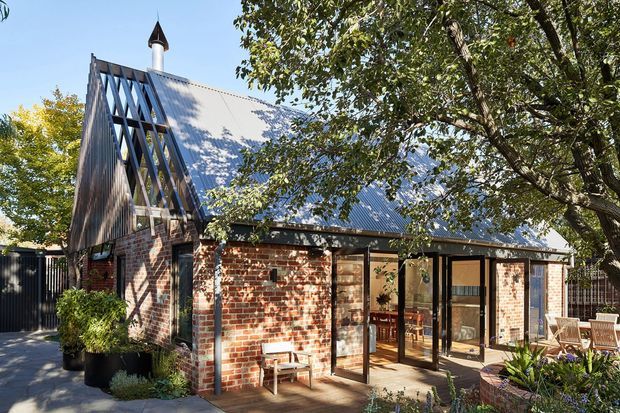 A compact brick building with a steep pitched gabled roof, dark timber or metal diagonal structural framing, and a small cupola-like roof vent. The facade combines exposed red brick, extensive glazed doors and windows, and a timber deck or paved outdoor sitting area. Mature trees partially frame the sunny exterior.