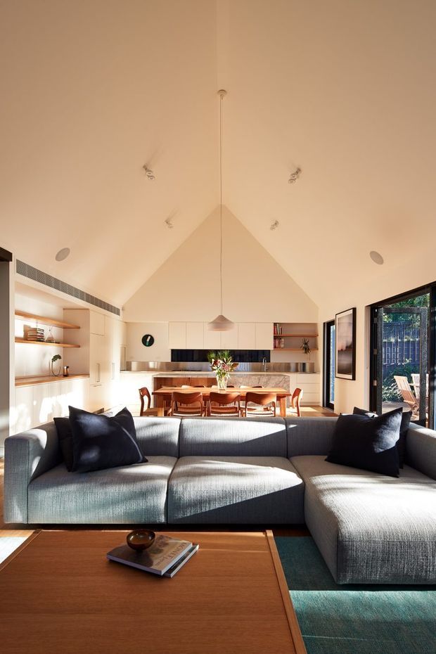 Bright open-plan living, dining and kitchen interior viewed from the living area. A large grey L-shaped sectional sofa faces the camera, with a timber coffee table in the foreground. Behind it is a timber dining table with upholstered chairs and a compact contemporary kitchen featuring light cabinetry, timber open shelving, a dark splashback and stone-look benchtop. The room has a dramatic vaulted ceiling with recessed spotlights and a central pendant, extensive glazing and sliding doors opening to an outdoor area. The palette is warm and refined, combining light neutral surfaces, timber and charcoal accents.