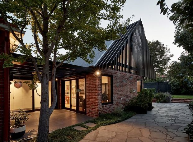 Exterior view of a contemporary brick dwelling or extension at dusk, featuring a sharply pitched gabled roof screened with closely spaced dark vertical battens. Full-height glazed doors and windows open onto a covered outdoor area, while a paved stone path and landscaped garden frame the building. Warm interior lighting contrasts with the dark timber-like facade screening and exposed brick walls.