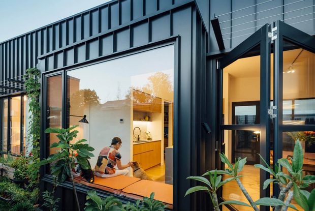 Exterior view of a contemporary dark-clad building with large glazed openings and folding glass doors. Warmly lit interior spaces are visible through the glazing, including a compact kitchen and living area, while dense plants soften the facade. The building has a strong rectilinear form and projecting roofline details.