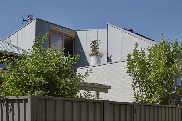 Exterior view of a contemporary multi-level residential building, partially screened by mature garden planting and a dark metal fence. The building features angular roof forms, pale fibre-cement or concrete-like wall panels, a white masonry lower wall, timber-framed glazing, and a corrugated metal roof section. Clear blue sky and strong daylight emphasize the clean geometric composition.