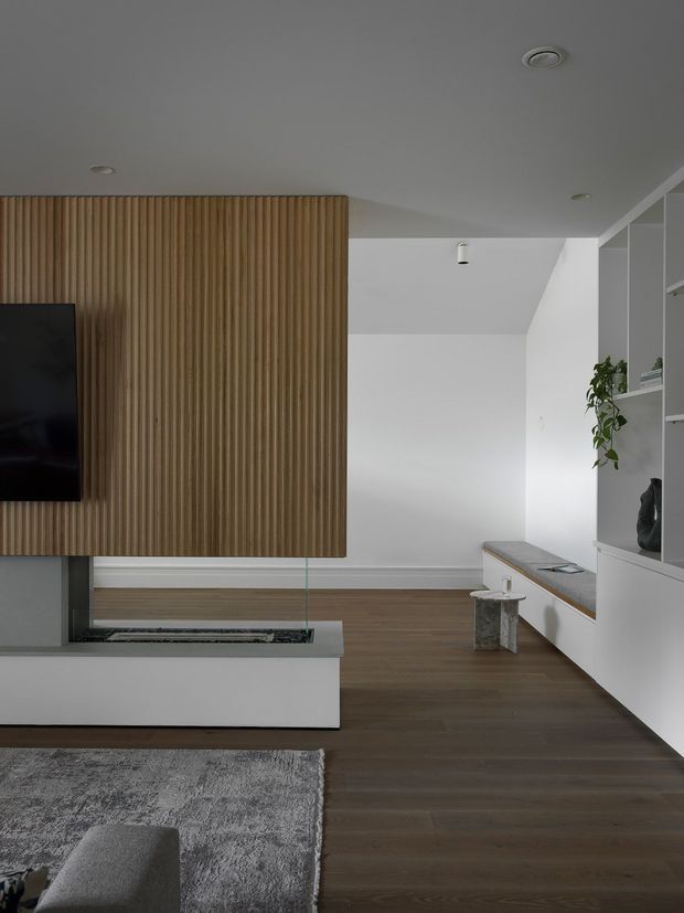 A contemporary living room with a suspended timber slatted feature wall incorporating a television and fireplace, white built-in cabinetry and shelving, a long window seat, dark timber flooring, and a large grey rug. The space has a restrained, minimalist composition with recessed and compact ceiling lighting.