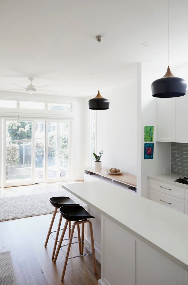 Bright, open-plan white kitchen with a large island and two black pendant lights over a timber-stool breakfast bar. White cabinetry and benchtops are paired with a dark grey tiled splashback, while full-height glazed doors open toward a sunlit outdoor area. Light timber flooring and restrained colourful artwork add warmth and contrast.