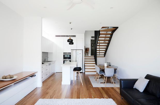 Bright open-plan living area with a compact white kitchen, central island and stools, dining table beneath an open timber staircase, and a black leather sofa in the foreground. The room features white walls and cabinetry, warm timber flooring, timber stair treads with dark framing, and abundant natural light.