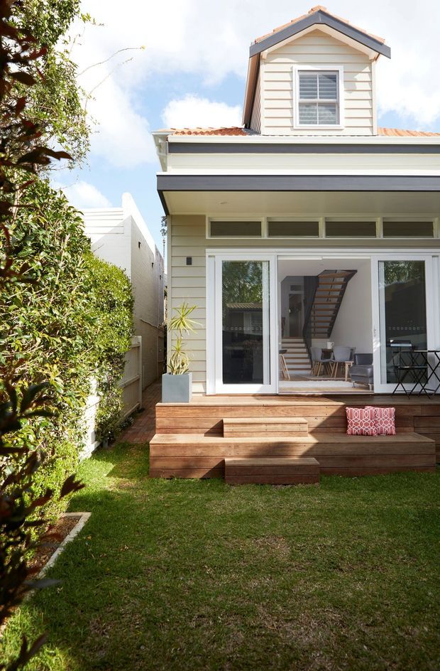 Bright exterior view of a narrow two-storey weatherboard house with a projecting gabled dormer, tiled roof, white trim, and large glazed doors opening onto a timber deck. The landscaped backyard includes lawn, hedges, and built-in timber bench seating; an interior staircase and living area are visible through the doors.