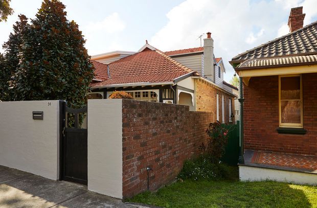 Street-level exterior view of a character residential property, showing a red brick boundary wall and gate, rendered front fence, tiled pitched roofs, chimneys, timber-framed windows, and a covered front porch with patterned tile flooring. The scene appears to be in an established suburban streetscape with traditional detailing.