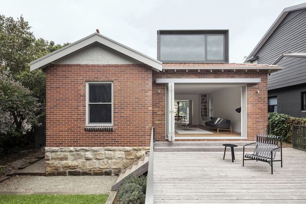 Exterior view of a compact brick house extension with a raised timber deck and open glazed connection to the living room. The original gabled brick volume has a light stone base and white roof trim, while the newer flat-roofed addition features clerestory-style upper glazing and wide opening doors. A simple outdoor seating area occupies the deck, with neighbouring residential architecture visible at the side.