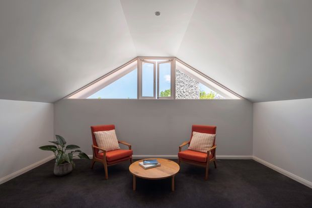 A compact attic-style sitting room with a sharply pitched white ceiling and a distinctive triangular clerestory window. Two mid-century-inspired red upholstered lounge chairs face a round timber coffee table, with a potted plant at the side. Dark carpet, white walls, and bright daylight create a simple, calm retreat.