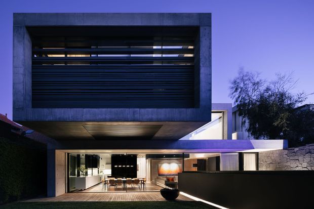 Twilight exterior view of a contemporary multi-level residence. A large cantilevered upper volume with dark horizontal louvres projects over a glazed ground-floor living and dining area. The facade combines smooth grey concrete, extensive glazing, dark screening, and a stone boundary wall, with warm interior lighting visible inside.