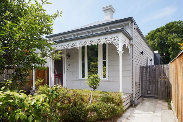 Street-facing view of a restored single-storey weatherboard cottage with a decorative Victorian-style verandah, ornate fretwork, turned posts, tall sash-like windows, a parapet-like roof detail, and a prominent chimney. The facade is painted white and surrounded by a compact landscaped garden, with a narrow paved side path and timber fencing.