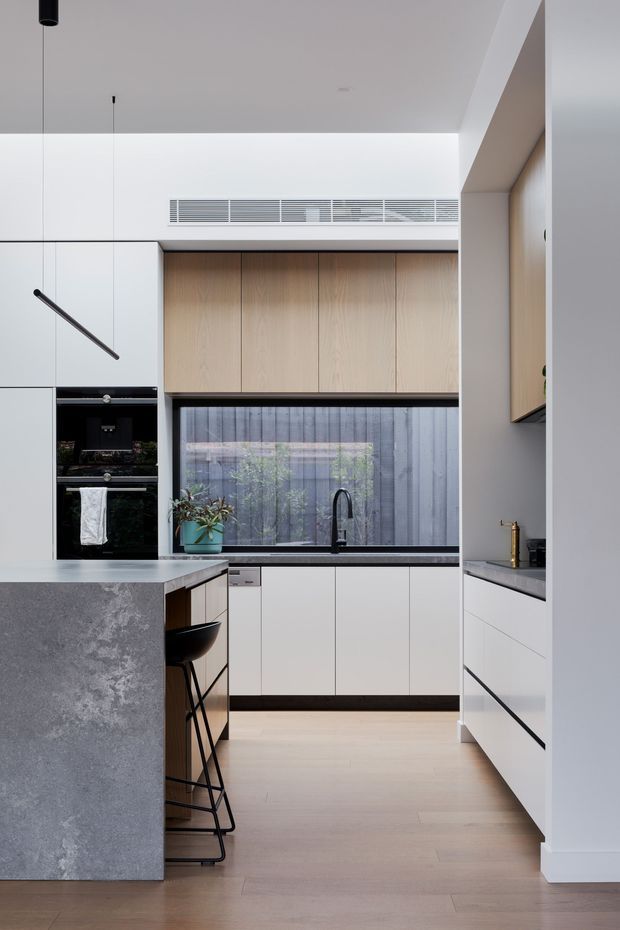 A contemporary galley-style kitchen with a central island, integrated black oven, white handleless cabinetry, warm timber overhead cupboards, dark stone surfaces, and a large window overlooking a planted courtyard. Minimal black pendant lighting, a black tap, and bar stools provide contrast against the pale walls and light timber floor.