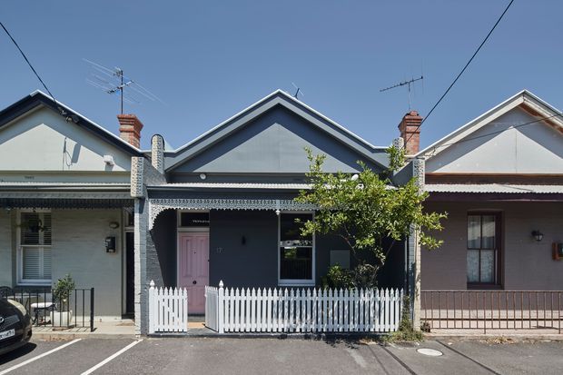 Street-facing row of attached heritage cottages with narrow front verandahs, decorative lacework, pitched gabled roofs, brick chimneys, painted masonry facades, timber-framed windows and small fenced front gardens. The central cottage is dark blue-grey with a pale pink door and white picket fence, flanked by lighter neighbouring cottages. Viewed from the street in bright daylight.