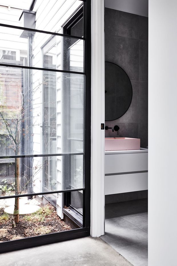 A contemporary bathroom viewed through a full-height black-framed glazed opening. The bathroom features dark grey large-format wall and floor tiles, a wall-mounted white vanity with integrated drawers, a pale pink vessel basin, matte black tapware, and a large round dark mirror. The glazing looks onto a small internal courtyard and brings abundant natural light into the space.
