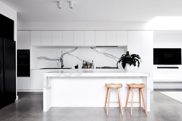A bright, minimalist open-plan kitchen with extensive white full-height cabinetry, a large white marble-look island, and a matching veined splashback. The island includes seating for two with natural timber stools. Dark polished concrete flooring, black appliances and fixtures, and a wall-mounted television create contrast, while a potted plant adds organic detail.