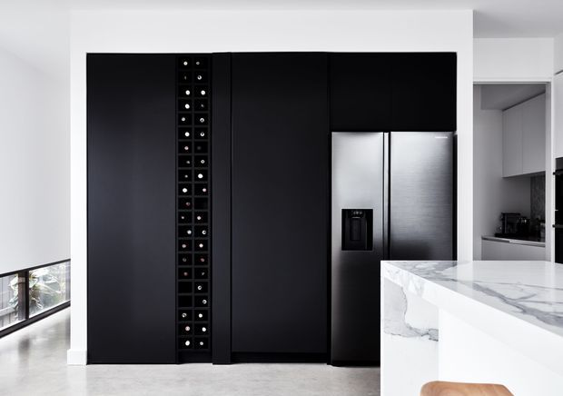 A contemporary kitchen viewed straight on, dominated by a tall matte-black wall of integrated cabinetry and a stainless-steel double-door refrigerator. A narrow built-in wine storage rack is set into the cabinetry. In the foreground, a large white marble-look island with pronounced grey veining and a waterfall edge is partially visible. White walls, pale flooring, and a glimpse of additional grey cabinetry create a crisp, high-contrast monochromatic scheme.