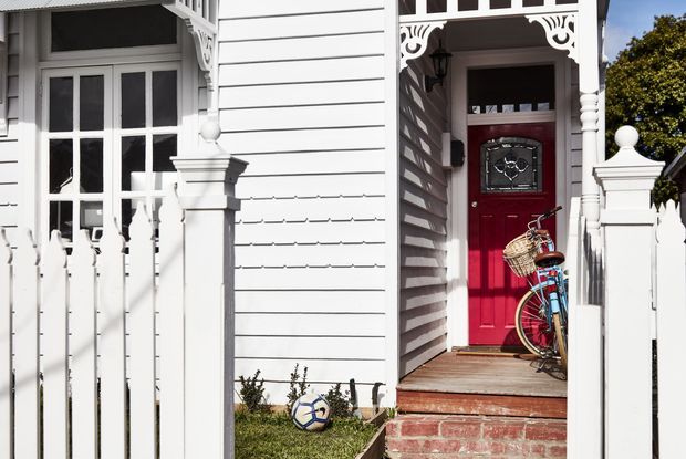 Sunlit exterior entry of a traditional weatherboard house, featuring white horizontal cladding, ornate timber fretwork, decorative porch posts, a red front door with glazed detailing, timber steps, and a white picket fence. A bicycle and soccer ball are present near the entrance.