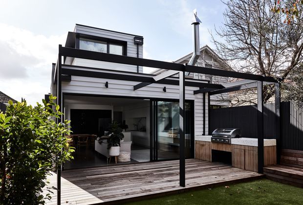 Rear exterior view of a contemporary multi-level home with a white horizontal weatherboard-clad extension, dark-framed glazing, and a timber deck. A black steel pergola or awning structure extends over the deck and outdoor kitchen, which includes a built-in barbecue and timber cabinetry. The open glazed doors reveal part of an interior living and dining area. The composition includes a dark privacy fence, steps, planted screening, and a visible pitched roof and chimney behind the extension.