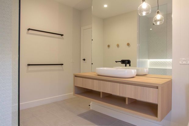 A contemporary bathroom with a floating timber vanity, white vessel basin, matte black tapware and wall-mounted towel rails. A large mirrored wall expands the room visually, with pendant lights suspended in front of it and a softly patterned glass shower screen visible at right. The room has pale neutral walls and large light-grey floor tiles.