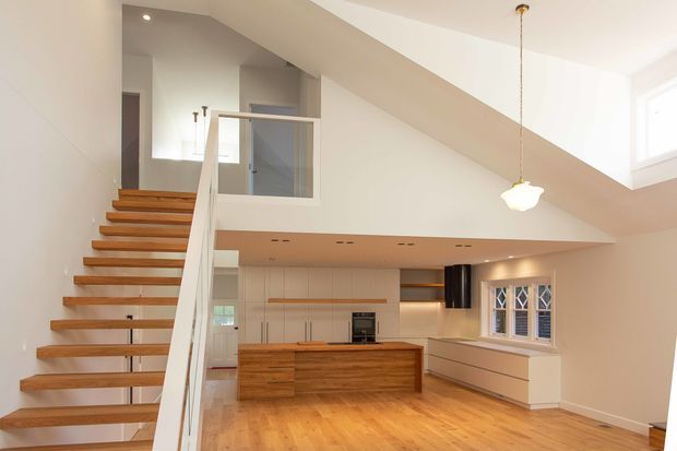 Bright open-plan interior featuring a timber staircase with white balustrade, a contemporary kitchen with extensive wood cabinetry and island, and warm timber flooring. The kitchen and living area sit beneath a dramatic sloping ceiling and open stair void, with clerestory-style windows, pendant lighting, and a built-in window seat or cabinetry.