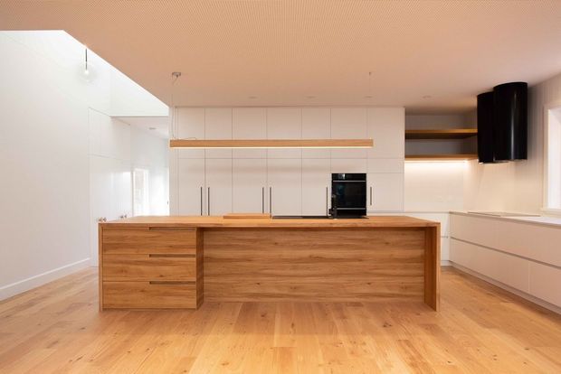 Bright, minimalist contemporary kitchen dominated by a large timber island with integrated drawers and a broad white built-in cabinetry wall. Light timber flooring, white surfaces, concealed appliances, open shelving, and a black cylindrical rangehood create a clean, warm-toned interior.