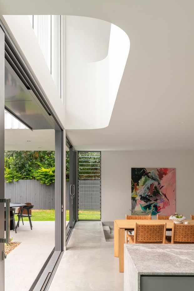Bright contemporary open-plan interior centered on a dining area and kitchen, with full-height glazed sliding doors opening to a covered outdoor patio. The space features a polished concrete floor, pale neutral walls, a sculptural curved white ceiling bulkhead, dark slender window frames, a timber dining table with woven timber chairs, and a large abstract artwork. A stone kitchen island occupies the foreground, while the outdoor dining area and landscaped courtyard are visible beyond the glazing.