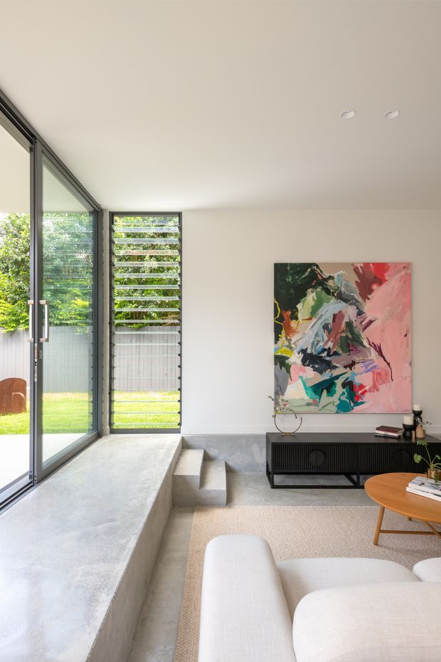 Bright contemporary living room with a minimalist concrete window bench and integrated steps beside full-height glazing. The room has pale walls and concrete flooring, dark slender window frames, a black ribbed media console, a large colourful abstract artwork, a light neutral rug, and a cream sectional sofa. A landscaped courtyard or garden is visible through the glazing.