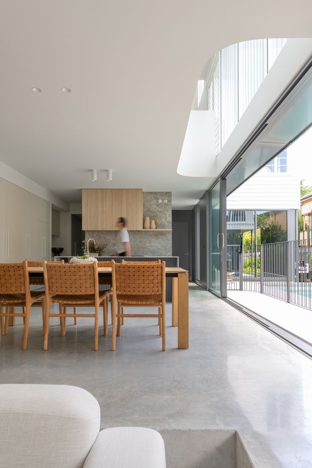A bright, open-plan contemporary kitchen and dining area with a timber dining table and woven timber chairs in the foreground. The kitchen features pale wood cabinetry, a dark stone-look splashback, and a central island. Full-height sliding glass doors open to an outdoor terrace or pool area, while a curved double-height opening and upper-level glazing bring in abundant natural light. A pale upholstered sofa and a sunken seating area are partially visible at the bottom of the frame.