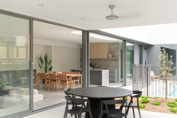 A bright contemporary indoor-outdoor entertaining area with a covered patio in the foreground, featuring a round dark dining table and chairs. Full-height sliding glass doors open toward an interior dining room and kitchen with timber cabinetry, a stone feature wall, and a marble-look island. A swimming pool and landscaped courtyard are visible beyond a slim metal balustrade. The space has a clean, refined, open-plan design with strong visual connection between the interiors and exterior.