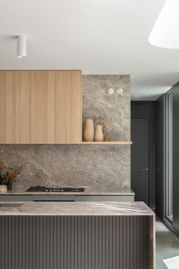 A refined contemporary kitchen featuring warm vertical timber cabinetry, a large grey-brown natural stone splashback and benchtops, and a dark fluted island front. A gas cooktop sits beneath the stone wall finish, with open shelving displaying ceramic vessels. Dark doors and vertical wall panelling continue into the adjoining area, while a large sculptural ceiling light and side glazing bring in soft natural light.