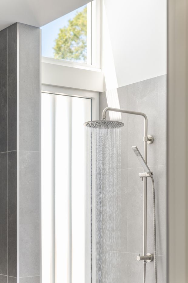 Bright, minimalist bathroom shower area with a large round rainfall showerhead and separate handheld shower on brushed silver fixtures. Light grey tiled walls, a darker grey tiled vertical feature wall, white surfaces, and tall frosted windows create an airy, contemporary interior.